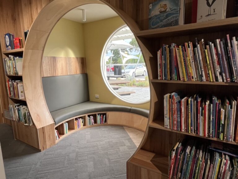 A cozy library nook features a circular wooden arch with a gray cushioned seat inside, surrounded by bookshelves filled with colorful books. A round window provides a view of the outside garden.