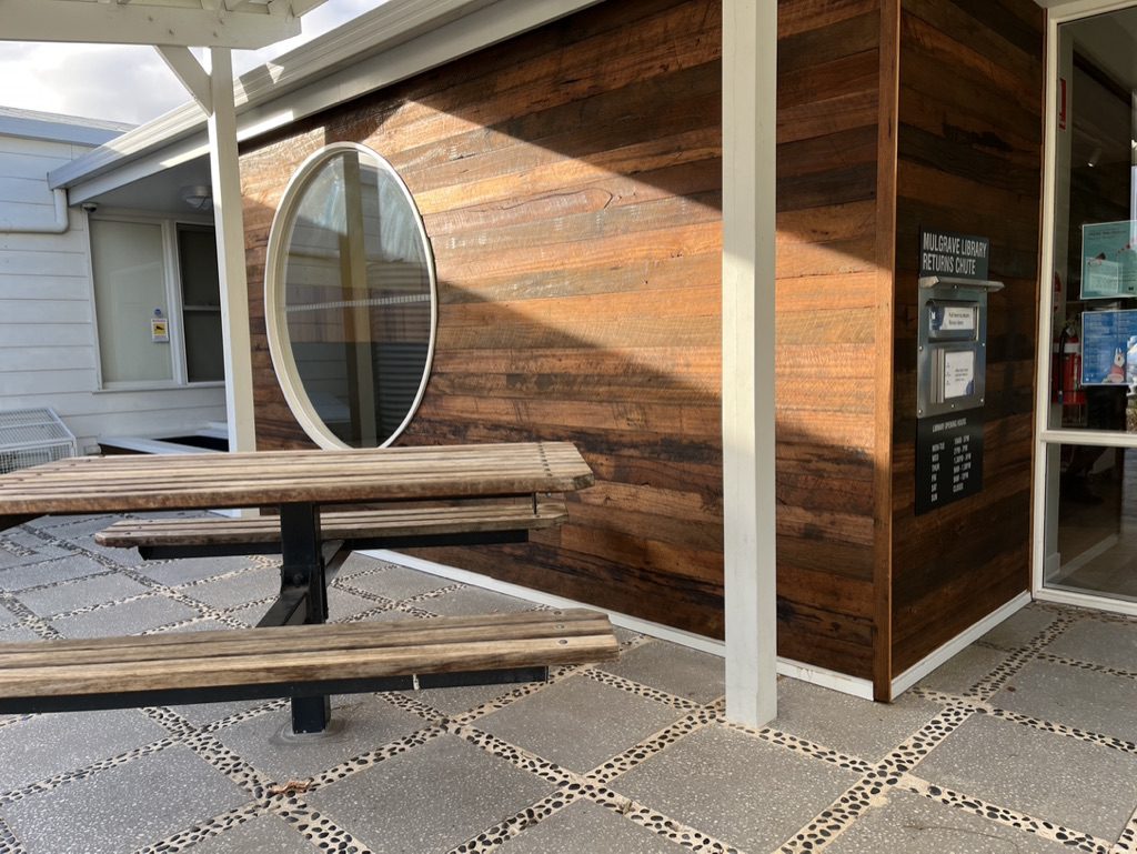 A wooden library entrance with an oval window and return slot is bathed in sunlight. A wooden picnic table is in the foreground, resting on a pebble and concrete patio.