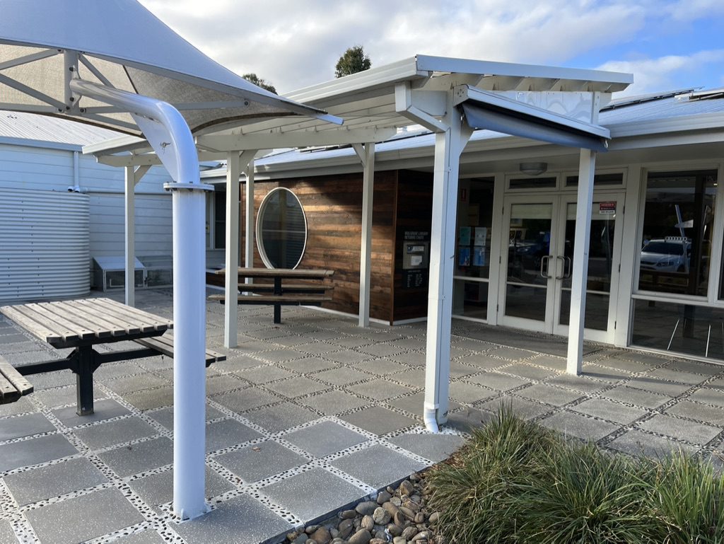 Outdoor area with a covered walkway and round window. Picnic tables are placed on a tiled patio. The building has a mix of wood and metal siding. Tall grass and a rocky border line the edge of the patio. Cloudy sky overhead.