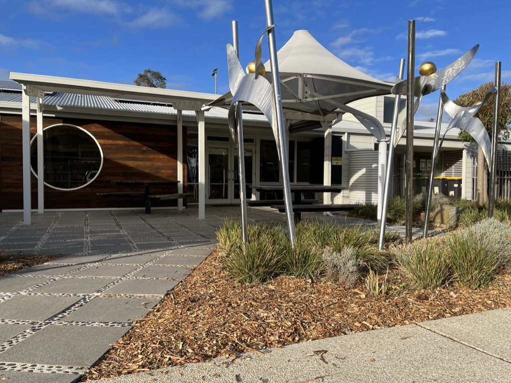 Modern building exterior with large circular windows, a geometric metal sculpture, and a landscaped garden in the foreground. The sky is blue with scattered clouds.