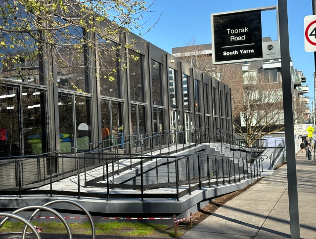 Street view of a modern glass-fronted building with multiple ramps and railings. A sign reads "Toorak Road South Yarra." Trees are in the background and there's a bike rack in the foreground. Clear blue sky above.