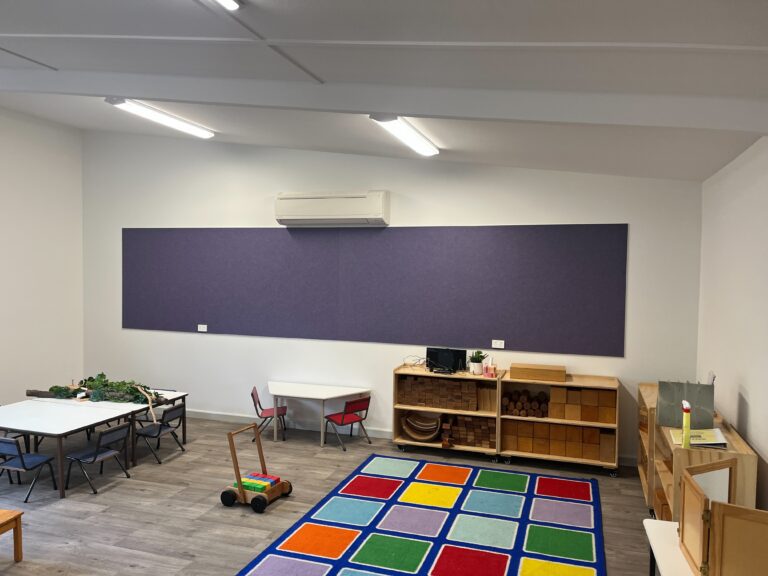 A classroom with small tables and chairs, a colorful square-patterned rug, wooden shelves, a toy push cart, and a wall-mounted air conditioner. The room is well-lit, with a minimalist design and open space.