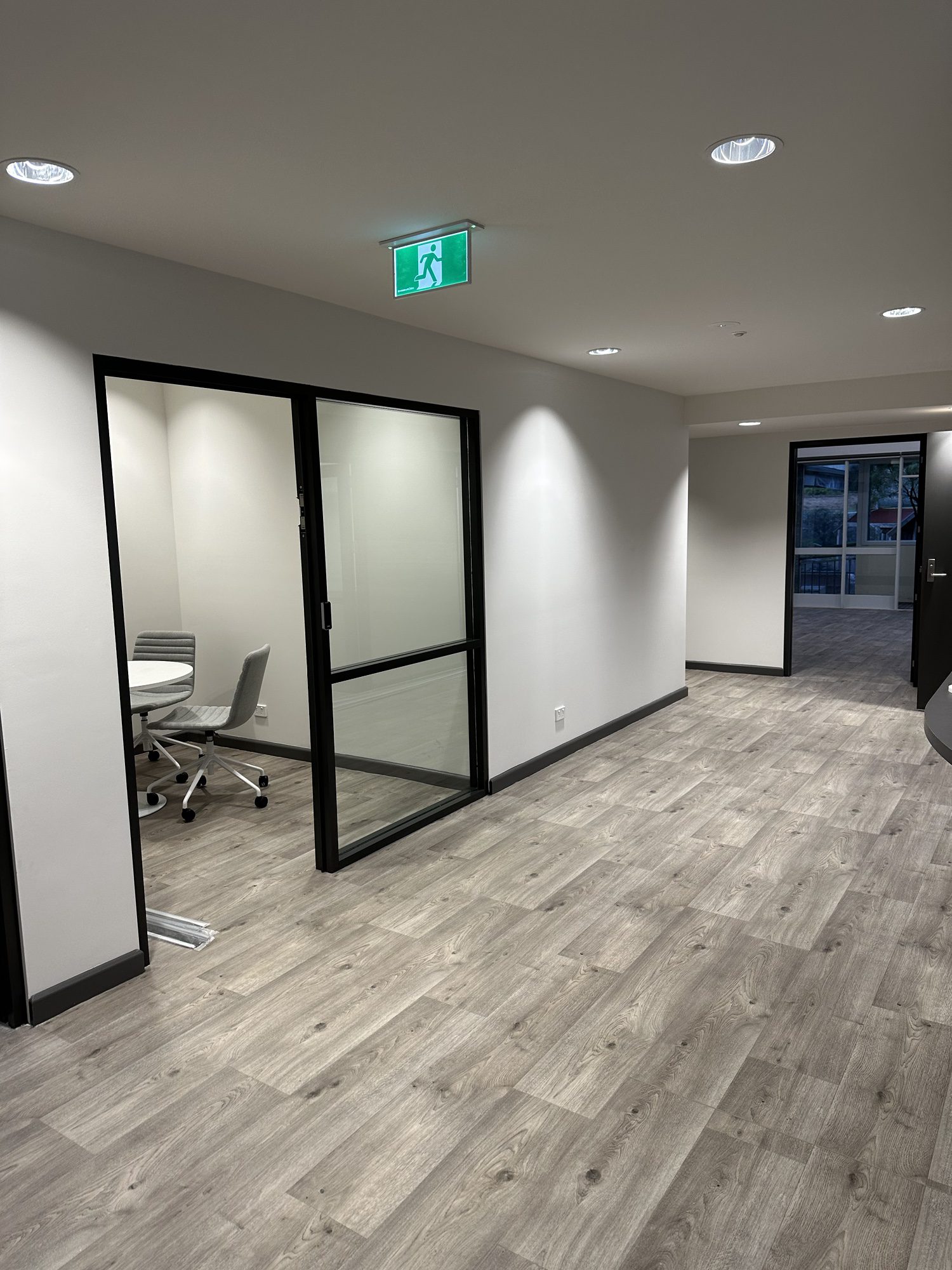 A modern office hallway with wooden flooring, white walls, and a small meeting room with two chairs visible through a glass door on the left. An illuminated exit sign hangs from the ceiling. A window at the far end reveals an external view.