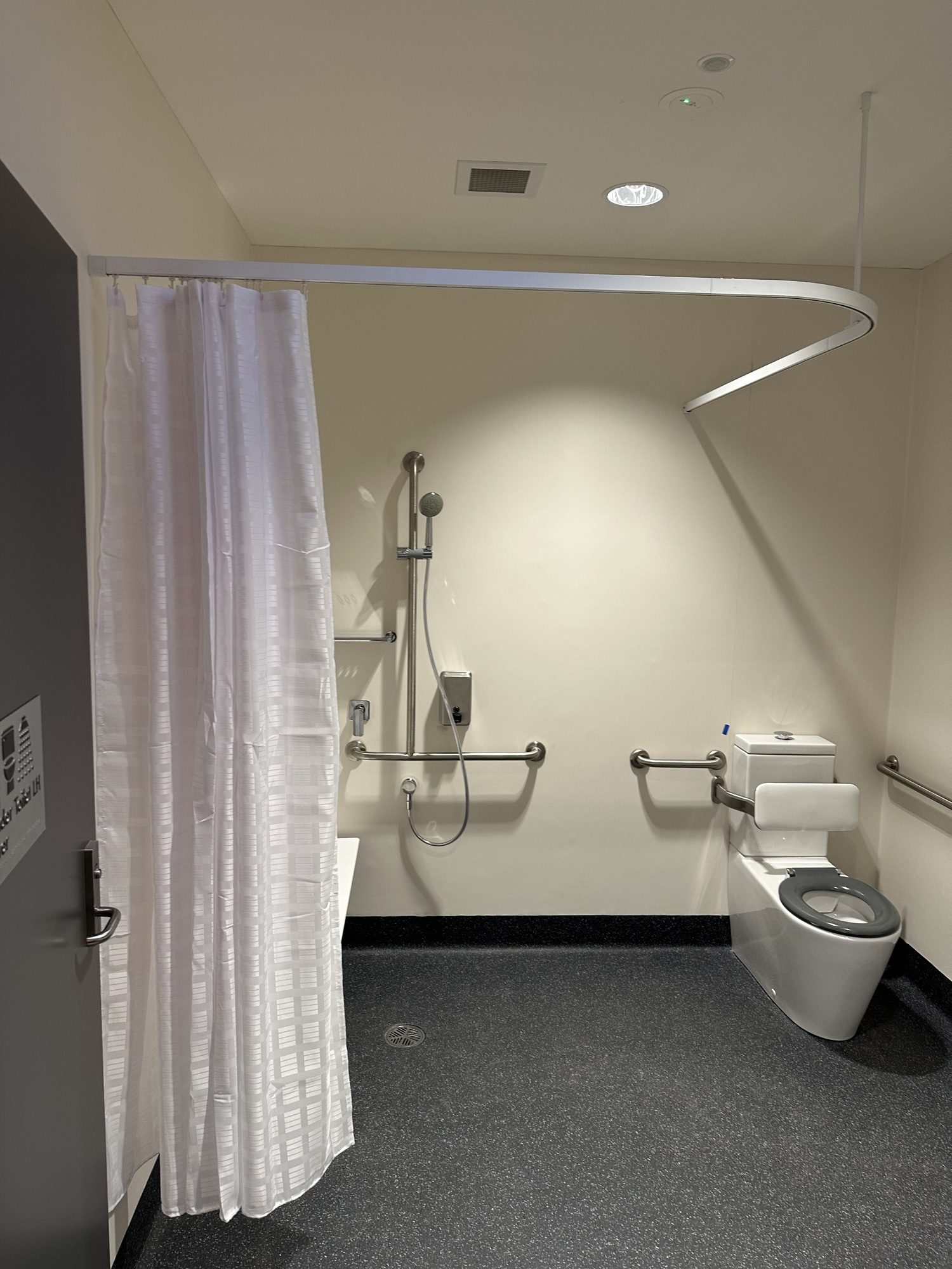 An accessible restroom with a white shower curtain, wall-mounted showerhead, grab bars, a toilet, and a sink. The floor is dark gray, and the walls are light-colored. Ceiling lights illuminate the space.