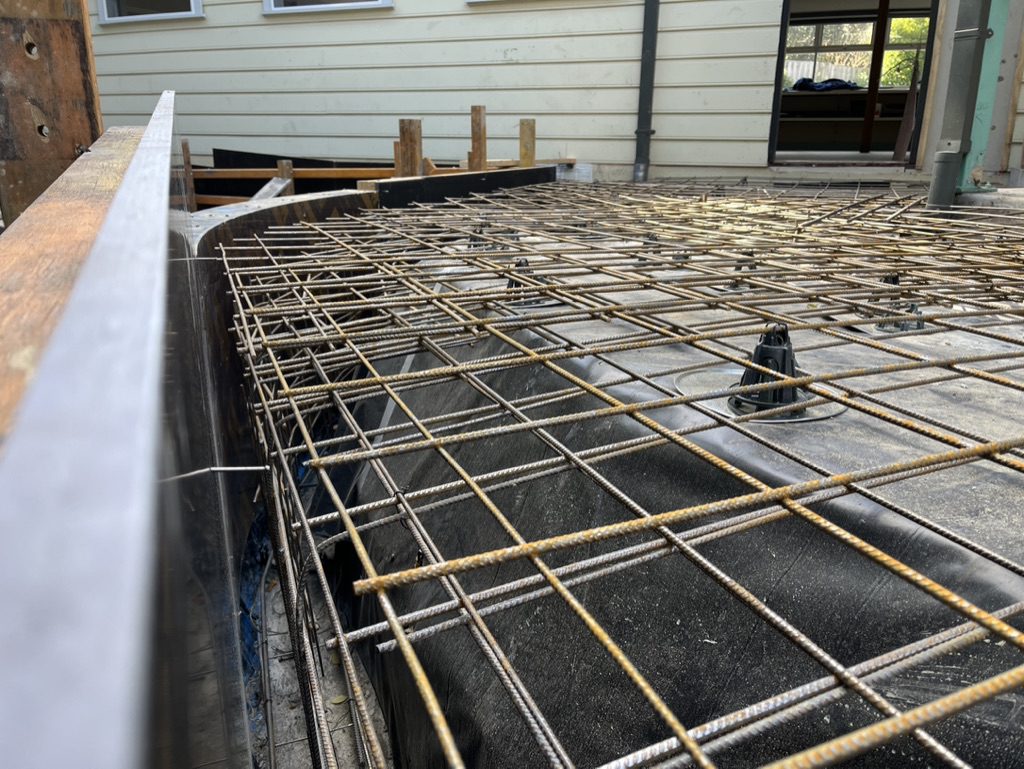 Close-up of a construction site showing a steel reinforcement grid atop a foundation, preparing for concrete pouring. The grid is framed by wooden planks, and a building is visible in the background.