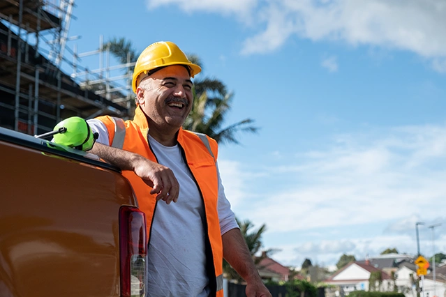 A construction worker in an orange vest and yellow hard hat leans against a truck, smiling. A safety helmet rests on the truck. The background features a building under construction and palm trees under a blue sky.