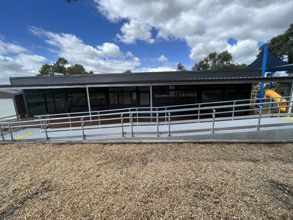 A building with large glass windows is fronted by a metal access ramp. A playground structure with a yellow slide is visible to the right. The sky is partly cloudy, and the ground is covered with wood chips.