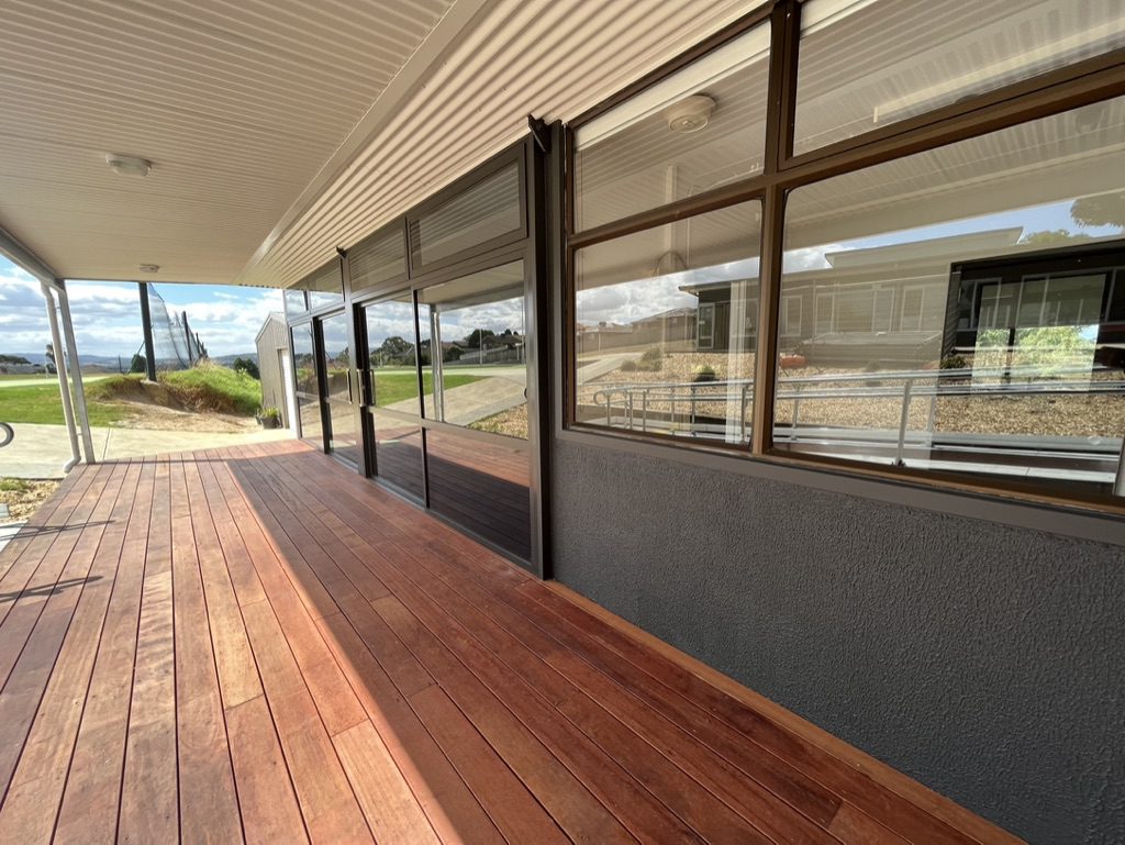 View of a wooden deck attached to a modern building with large windows. The area is covered by a slanted roof. In the background, there are paved pathways and various buildings under a partly cloudy sky.