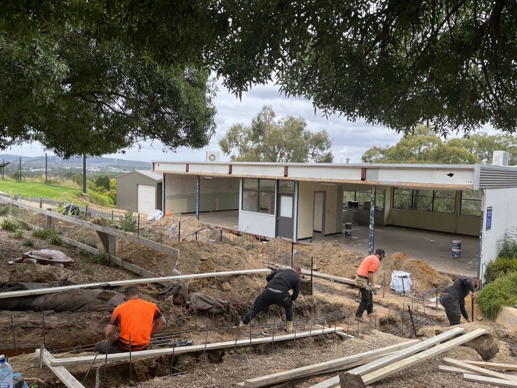 Four construction workers in safety gear work on a house extension outdoors. The existing building is white with large windows. The scene is surrounded by trees, and the sky is overcast. Building materials and tools are scattered around the site.