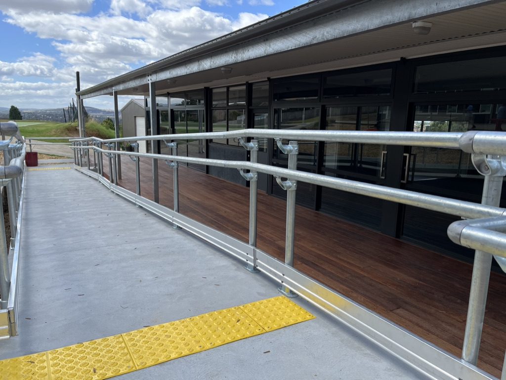 A long, metal accessibility ramp with yellow tactile paving leads to a building with large glass windows and a wooden deck. The sky is partly cloudy, and the landscape is grassy and open.