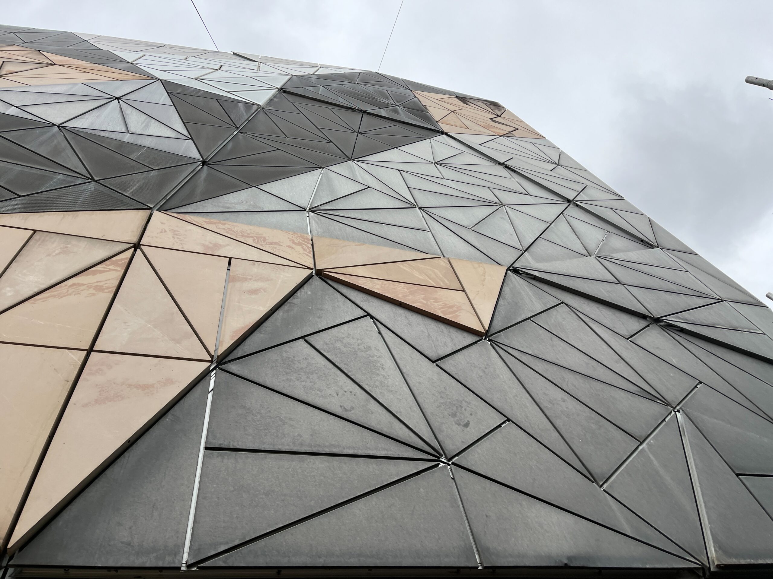 Geometric architectural structure with intersecting triangular panels in shades of gray and beige against a cloudy sky.