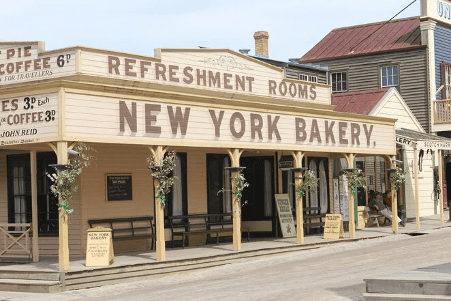 A wooden storefront with a sign reading "NEW YORK BAKERY" and "REFRESHMENT ROOMS." The building has a covered porch, decorated columns, and old-fashioned signs listing prices for pies and coffee.