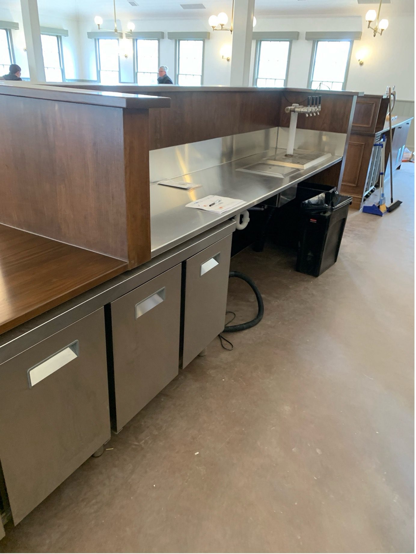 A commercial bar counter with stainless steel surfaces, multiple beer taps, refrigerators below, and wooden panels, set in a brightly lit room with windows and wall lamps.