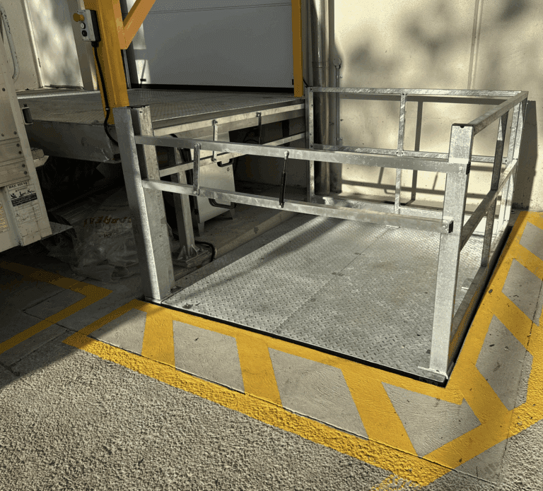 A metal loading platform with railings is positioned next to a truck at a loading dock. The ground is marked with yellow diagonal lines, designating a safety zone around the platform.