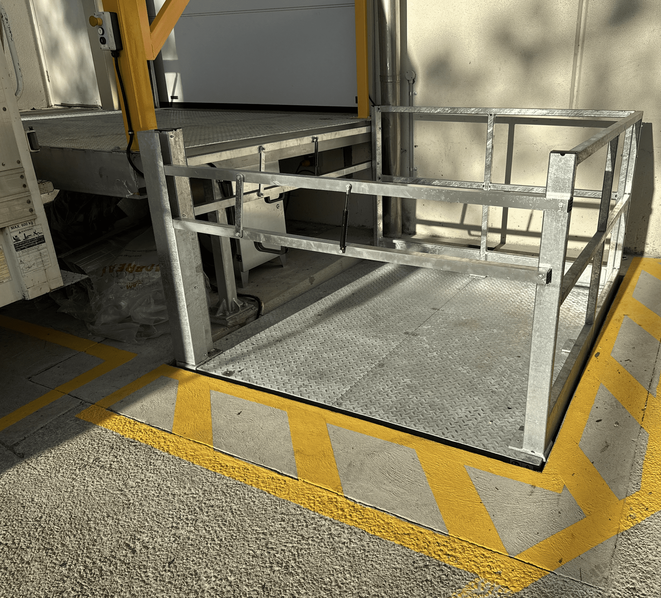 A metal loading platform with railings is positioned next to a truck at a loading dock. The ground is marked with yellow diagonal lines, designating a safety zone around the platform.