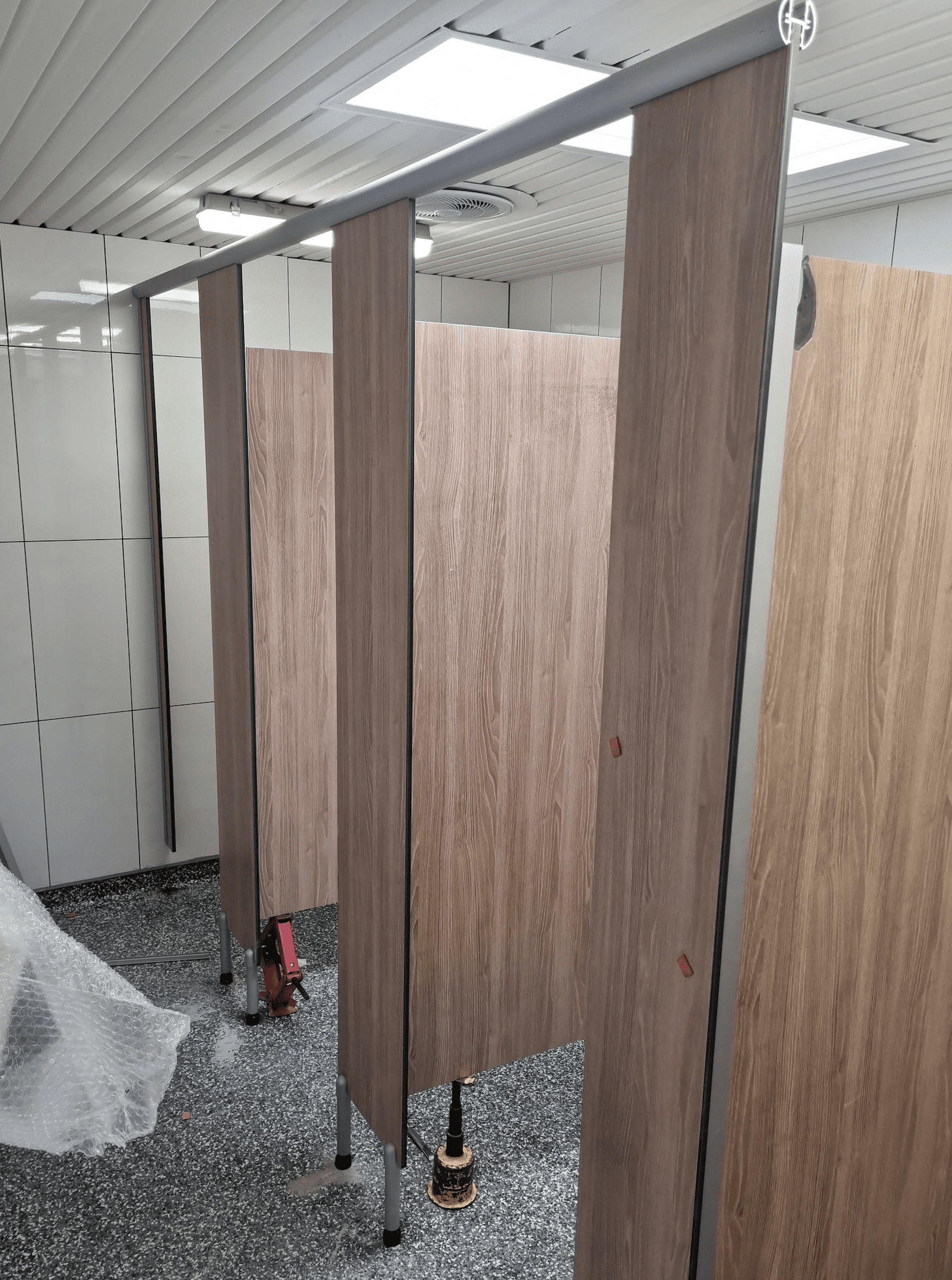 Three wooden restroom stall doors with metal frames are partially installed in a tiled bathroom. Tools and packing materials are on the floor, and ceiling lights are visible above.